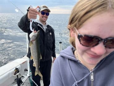 A proud angler holding a large Lake Trout while fishing on a boat, with a young companion smiling in the foreground.