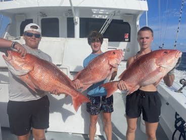 Three young anglers proudly displaying their catch of Red Snapper aboard a fishing charter in open waters.