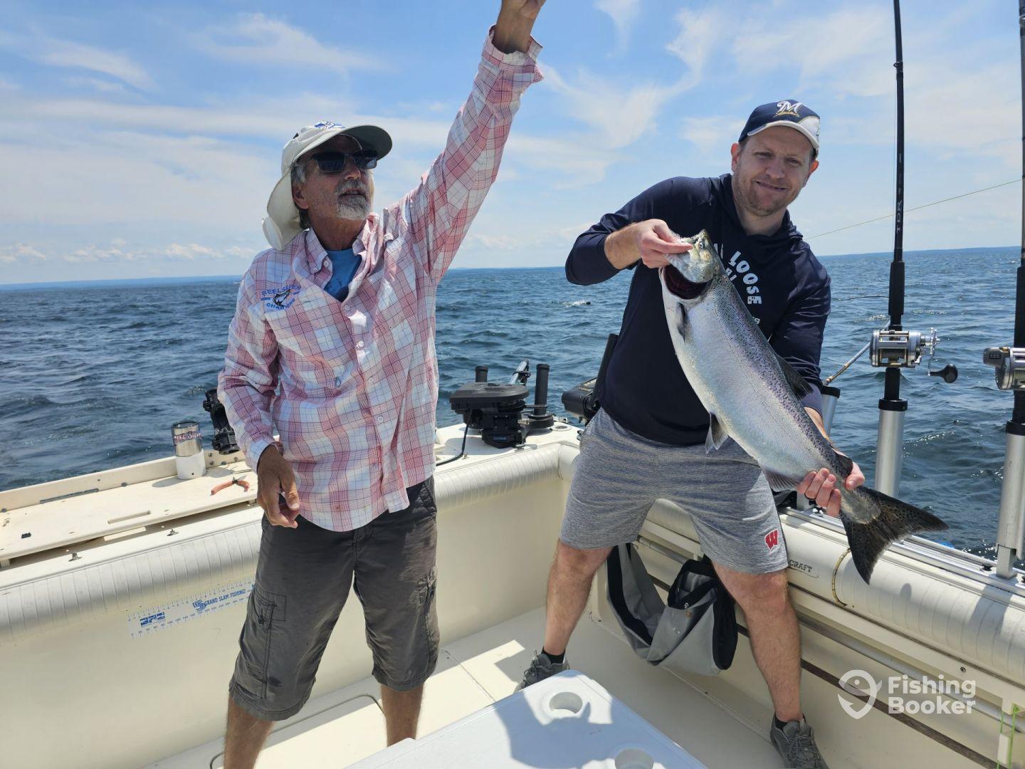Two anglers aboard a boat, one proudly displaying a large Salmon while the other gestures in excitement during a fishing trip.