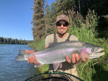 Angler proudly displaying a large Rainbow Trout caught in a scenic river setting surrounded by lush greenery.