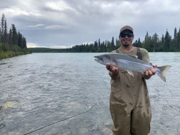 An angler proudly displaying a Silver Salmon caught in a scenic river setting, surrounded by lush greenery.