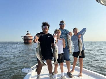 A family enjoying a successful fishing trip, proudly displaying their catch of striped bass on a boat near a lighthouse.
