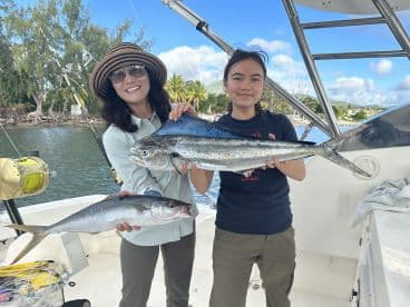 Two anglers, a woman and a young girl, proudly displaying their catch of Mahi Mahi and another fish aboard a fishing boat in a tropical setting.