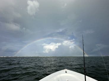 A scenic view from the bow of a boat, showcasing a rainbow over the water, with fishing gear visible in the foreground.