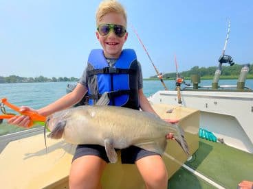 A young angler proudly displaying a large catfish while seated on a boat, enjoying a sunny day on the water.