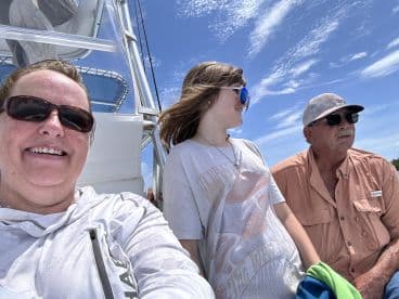 A family enjoying a sunny day on a fishing boat, with a scenic backdrop of blue skies and clouds.