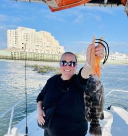 Angler proudly displaying a large Black Grouper while aboard a fishing boat, with a scenic waterfront in the background.