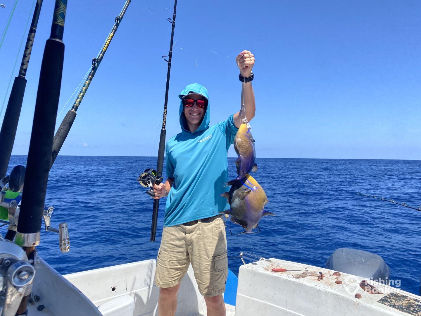 An enthusiastic angler proudly displaying two colorful fish, likely from a deep-sea fishing trip, aboard a boat in open water.