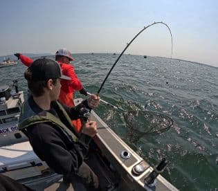 A young angler is actively reeling in a fish while another angler assists with a net on a fishing boat, showcasing the excitement of fishing in open waters.