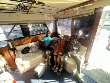 Three individuals relaxing inside a spacious cabin of a fishing boat, showcasing comfortable seating and a view of the marina through large windows.
