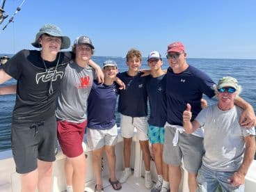 A group of young anglers and an adult enjoying a day out on the water, posing together on the boat with a clear blue sky in the background.