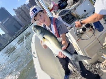 A young angler proudly displaying a Bluefish while fishing in an urban setting, with skyscrapers in the background.