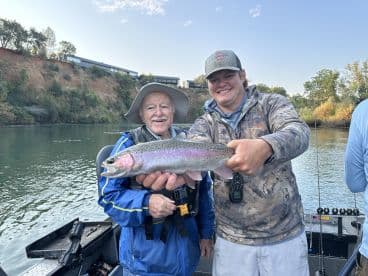 Two anglers proudly displaying a Rainbow Trout while fishing on a scenic river.