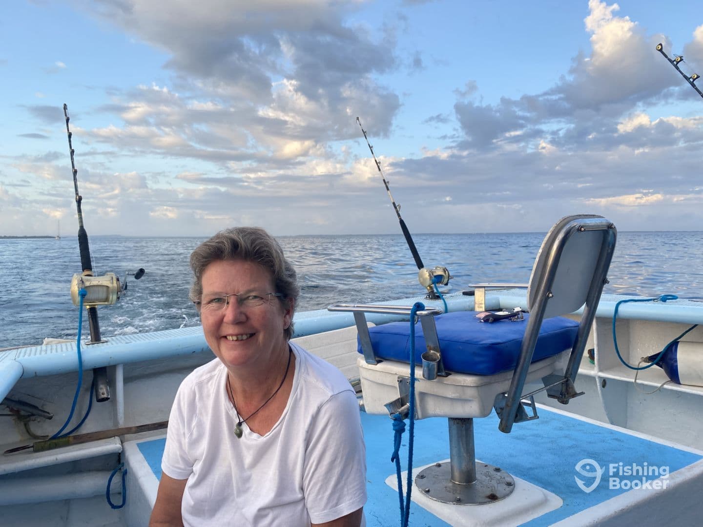 A smiling woman sits in the helm area of a fishing boat, enjoying a peaceful day on the water with fishing rods in the background.