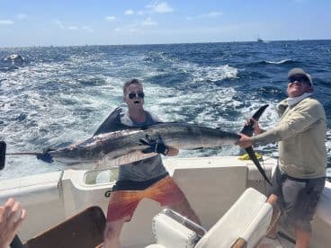 Two anglers proudly displaying a large Marlin while on a sport fishing boat in open waters.