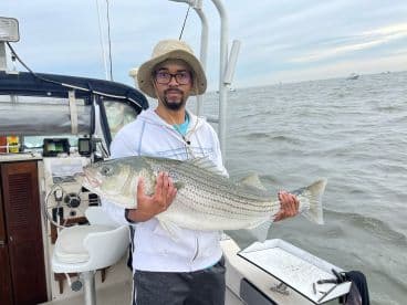 An angler proudly displaying a large Striped Bass aboard a fishing boat, showcasing a successful day on the water.