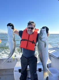 An angler proudly displaying two large Striped Bass while fishing on a boat, showcasing a successful day on the water.