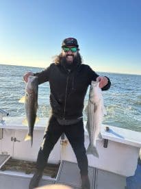 Angler proudly displaying two large Striped Bass while fishing on a boat, showcasing a successful day on the water.