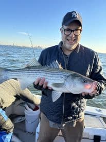An angler proudly displaying a large Striped Bass while fishing on a boat, with a scenic waterfront in the background.