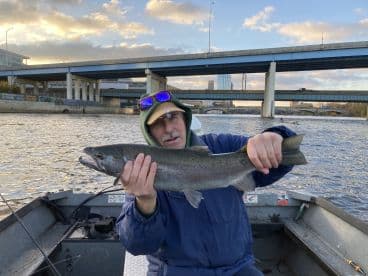 An angler proudly displaying a large Rainbow Trout while fishing from a small boat in a river setting, with urban scenery in the background.