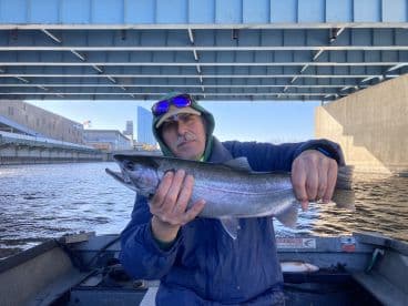 Angler proudly displaying a large Steelhead trout while fishing under a bridge in an urban setting.
