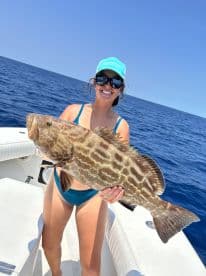 A smiling angler proudly displays a large Grouper while fishing offshore, showcasing a vibrant blue ocean backdrop.