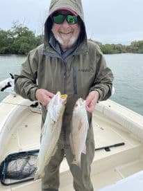 An angler proudly displaying two freshly caught fish, a Speckled Trout and a Redfish, while on a fishing boat in a coastal environment.