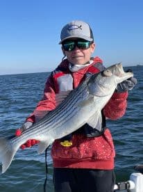 A young angler proudly displaying a large Striped Bass while fishing on a boat, showcasing a successful day on the water.