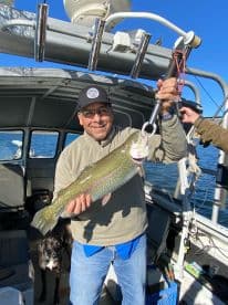 An angler proudly displaying a large Rainbow Trout aboard a fishing boat, enjoying a sunny day on the water.