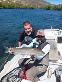 Two anglers proudly displaying a large Rainbow Trout while fishing on a scenic river.
