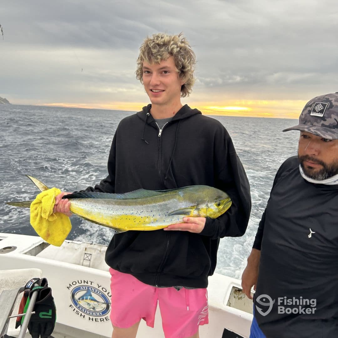 A young angler proudly displaying a vibrant Mahi Mahi on a fishing charter, with a scenic ocean backdrop at sunset.