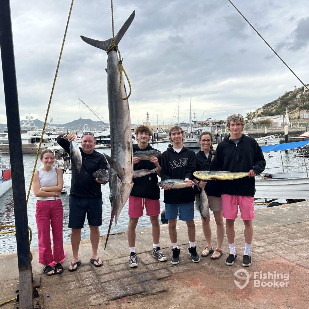 A family proudly displaying their catch, including a large Marlin and several smaller fish, at the dock after a successful fishing trip.