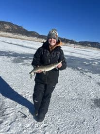 A young angler proudly displaying a Northern Pike caught while ice fishing in a scenic mountain lake.