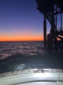 A serene sunset view from the side of a boat near an offshore platform, showcasing the calm waters and vibrant colors of the evening sky.