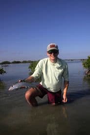 Young angler proudly holding a Snook while wading in shallow waters, surrounded by mangroves on a sunny day.