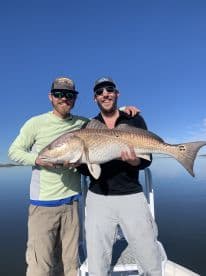 Two anglers proudly displaying a large Redfish while fishing on calm waters, showcasing a successful day on the boat.