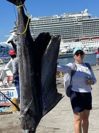 An angler proudly displaying a large Sailfish at the dock, with a cruise ship in the background, showcasing a successful fishing trip.