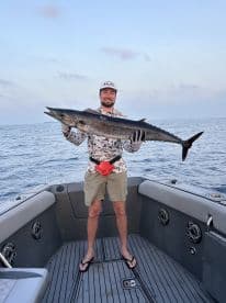 Angler proudly displaying a large Wahoo on the deck of a modern fishing boat during a deep-sea fishing trip.