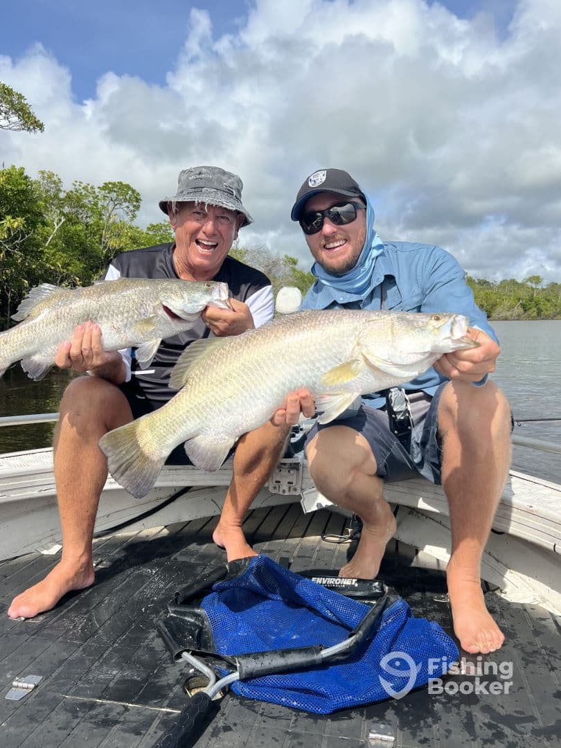 Two anglers proudly displaying their large Barramundi catches while fishing in a scenic river environment.
