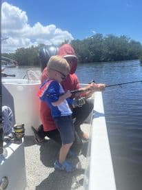 A young boy in a shark-themed shirt is learning to fish with an adult on a boat, enjoying a sunny day on the water.
