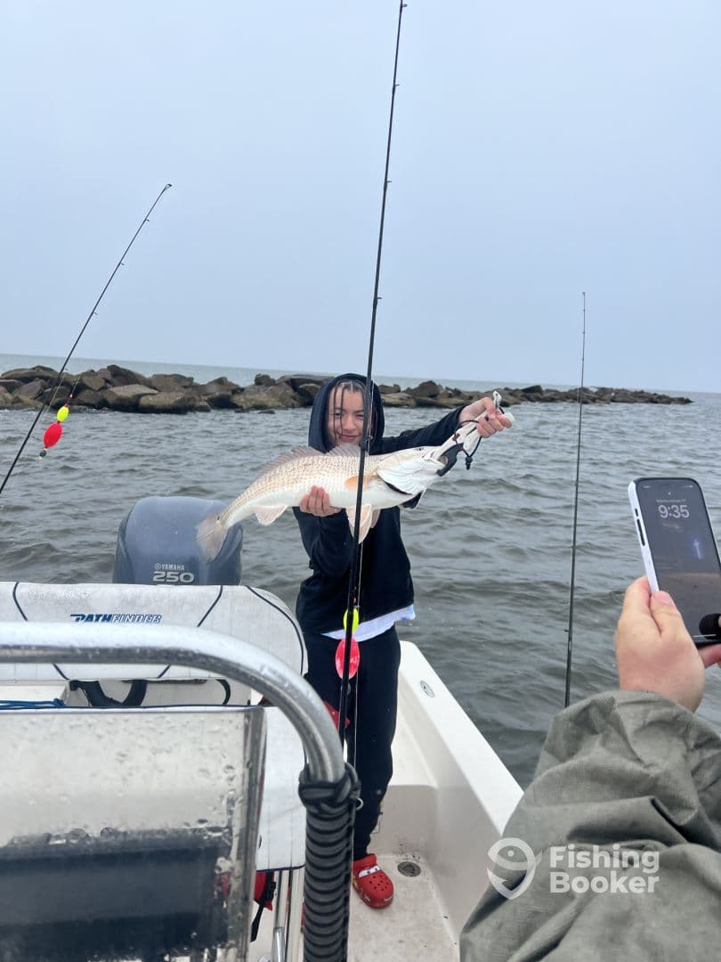 A young angler proudly displaying a large Redfish while fishing from a boat, with rocky formations in the background.