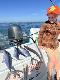 A young angler sits on the boat beside a catch of colorful fish, including Mackerel, showcasing a successful day of fishing in clear waters.