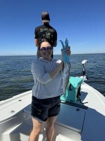 Angler proudly displaying a Speckled Trout while fishing on a sunny day, with a scenic water backdrop.
