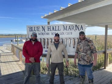 Three anglers proudly posing in front of a catch board displaying several Speckled Trout at Blue N Hall Marina, showcasing a successful day of fishing.