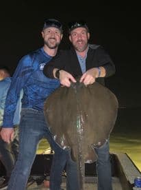 Two anglers proudly displaying a large stingray caught during a night fishing trip.