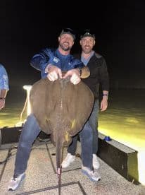 Two anglers proudly displaying a large stingray caught during a night fishing trip, showcasing the excitement of their catch.