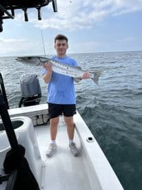 A young angler proudly displaying a Barracuda while standing on the deck of a fishing boat in open waters.