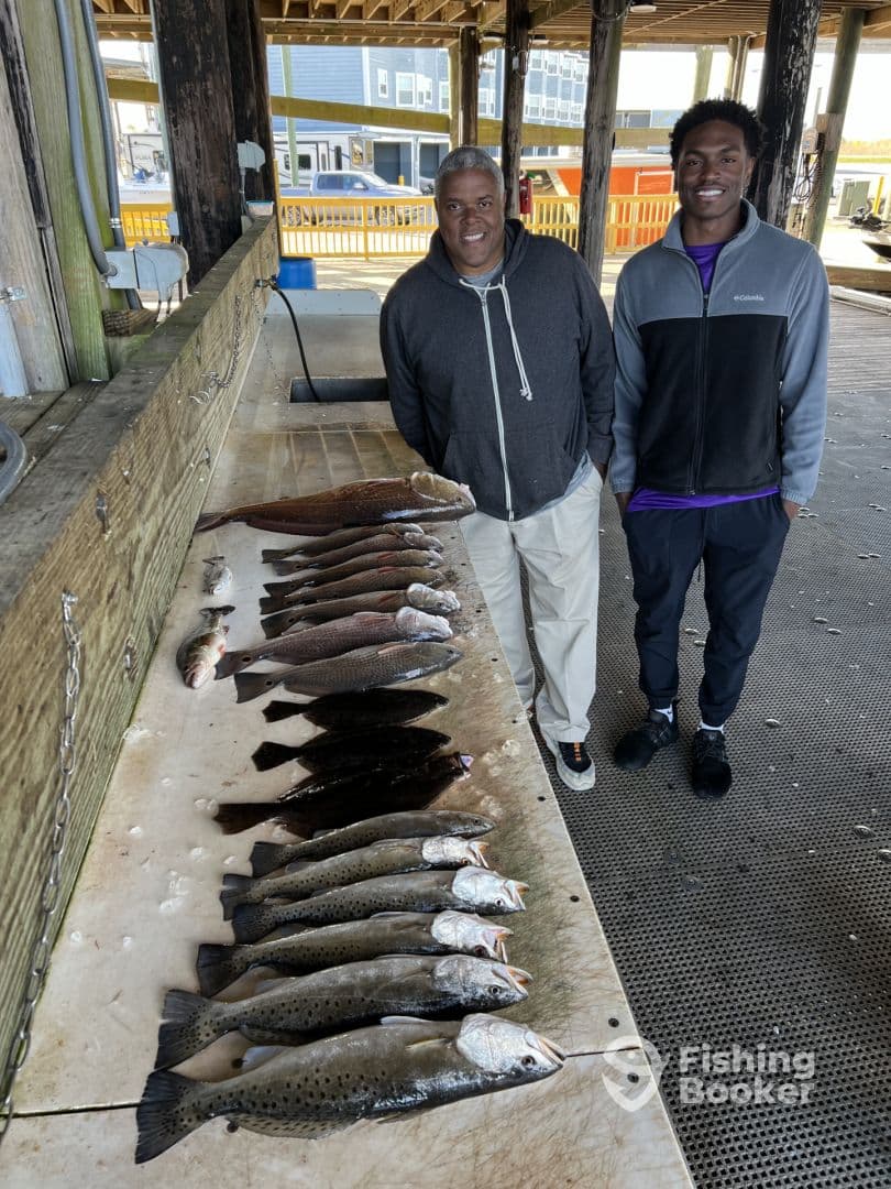 Two anglers proudly displaying a variety of fish, including Speckled Trout, arranged on a cleaning table at a fishing dock.