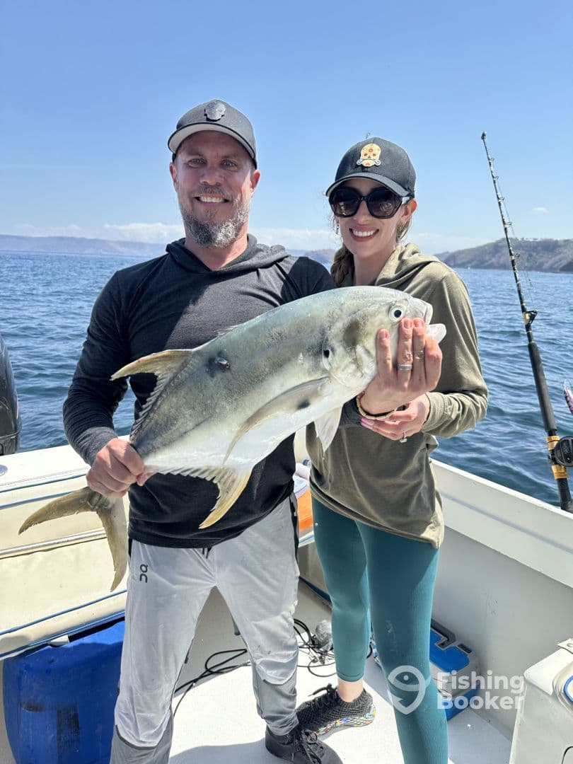 Two anglers proudly displaying a large Yellowtail while fishing on a boat in a sunny coastal environment.