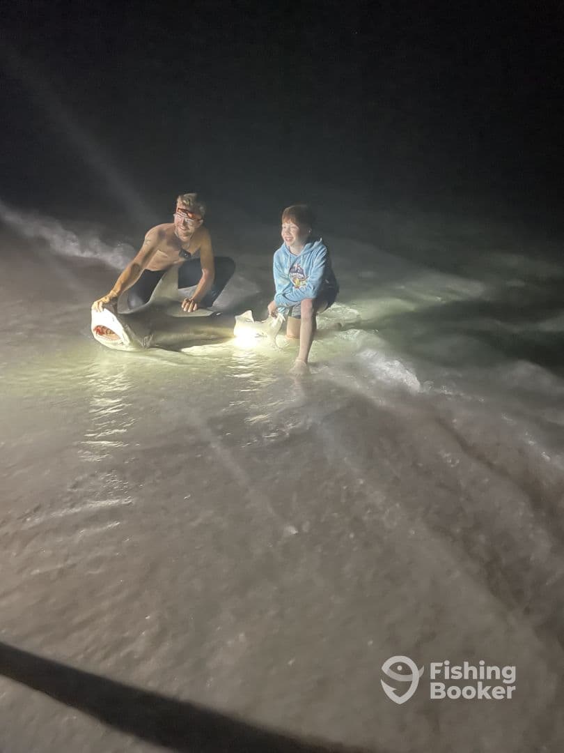 A young boy and an adult are joyfully posing with a caught shark on the beach at night, showcasing their fishing adventure.